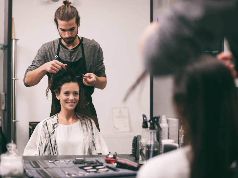 A close-up of a salon professional expertly working on a client's hair, visible through the client's satisfied reflection in the mirror.